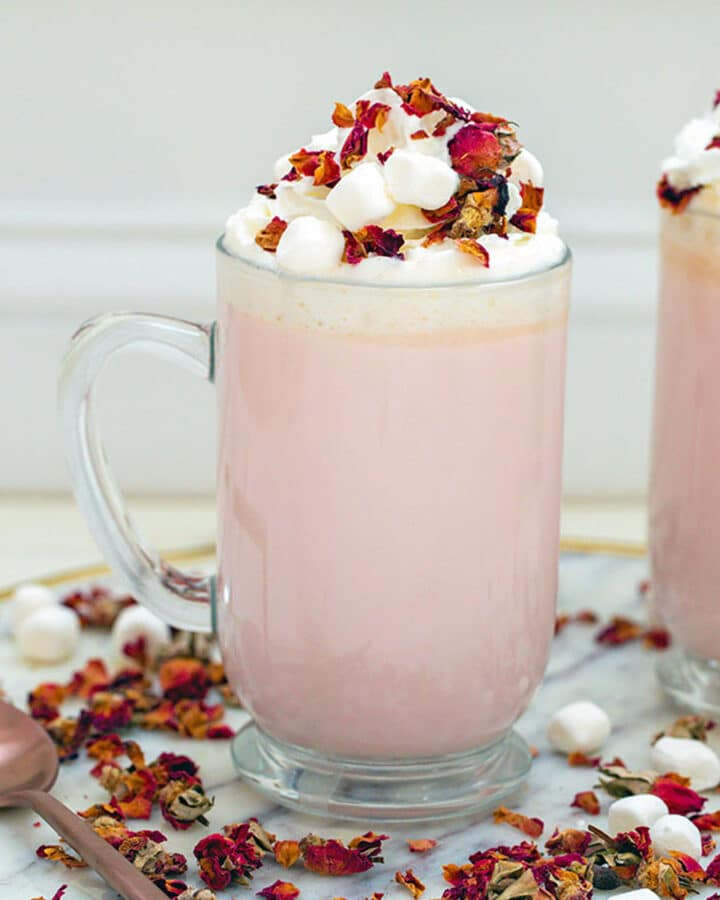 Head-on view of a large mug of pink colored rose white hot chocolate topped with whipped cream and rose petals with rose petals all around