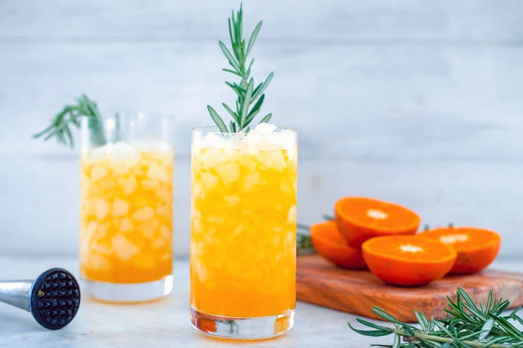 Landscape view of two tall glasses of tangerine and rosemary cocktail with rosemary garnish with cutting board with tangerines and rosemary and muddler in the background