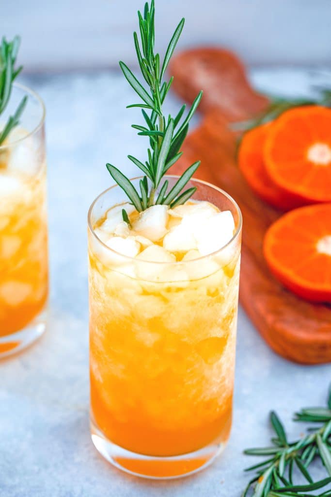 Close-up image of tangerine and rosemary cocktail in a tall glass with crushed ice and rosemary garnish with cutting board with tangerines and rosemary in the background