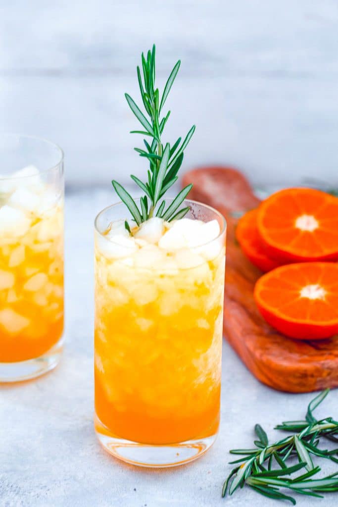 Head-on view of tangerine and rosemary cocktail in a tall glass with rosemary garnish and second cocktail, cutting board with sliced tangerines and rosemary in the background