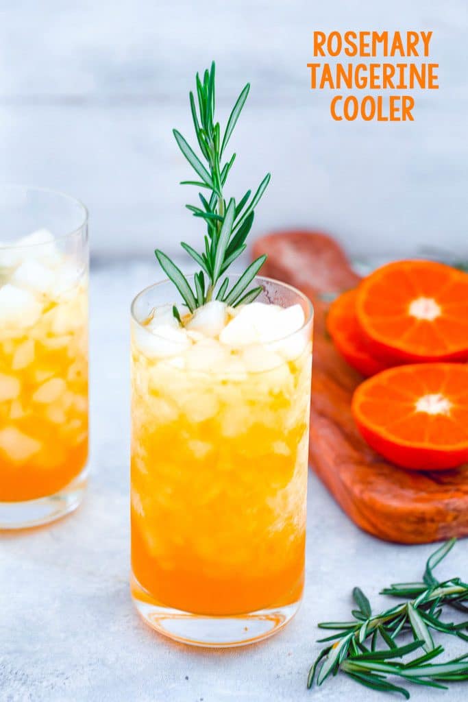 Head-on view of tangerine and rosemary cocktail in a tall glass with rosemary garnish and second cocktail, cutting board with sliced tangerines and rosemary in the background and recipe title at the top of the image