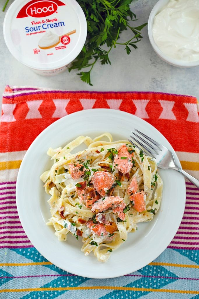 Bird's eye view of white plate with cream-covered fettuccine, salmon, bacon, and herbs with a fork and sour cream container, parsley, and bowl of caesar dressing in the background
