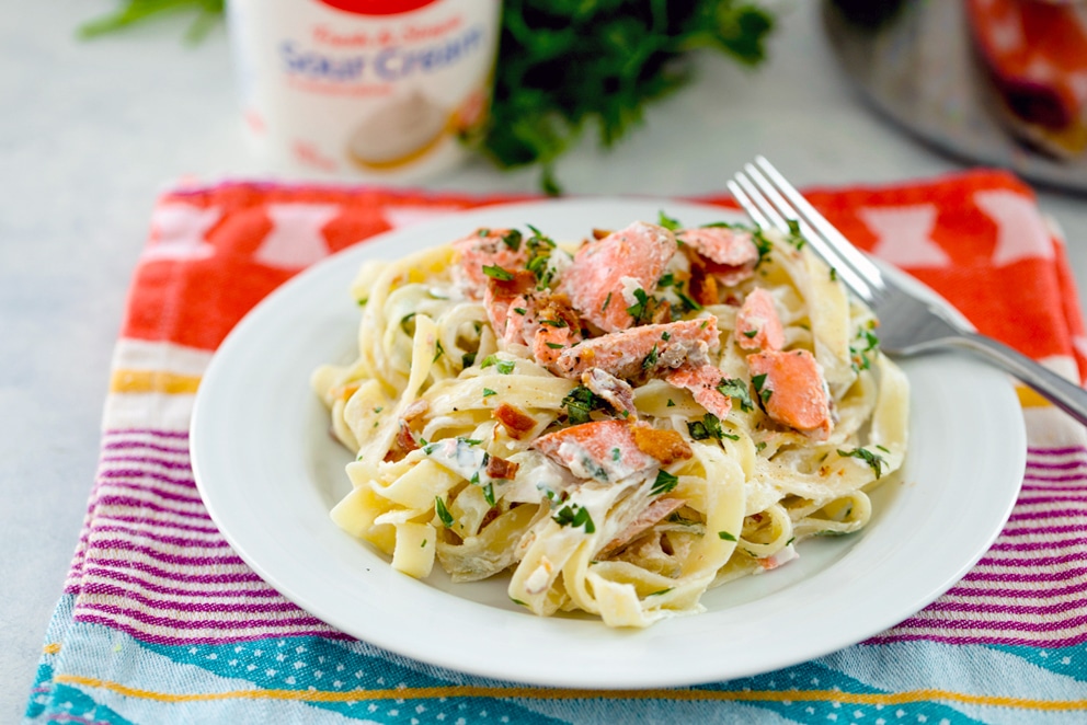 Landscape photo of white plate with cream-covered fettuccine with salmon, bacon, and herbs with fork on the plate and sour cream container and parsley in the background