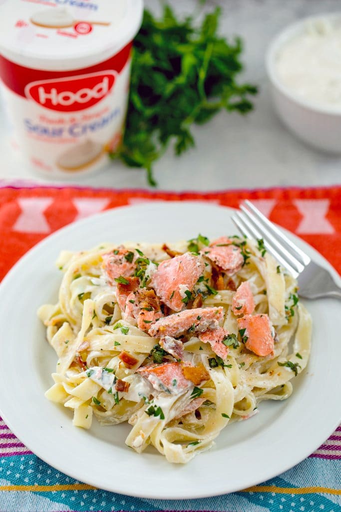 Creamy fettuccine on a white plate with salmon, bacon, and herbs and sour cream container and parsley in the background