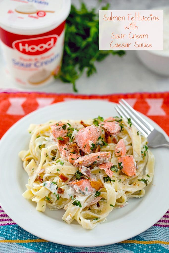 Creamy fettuccine on a white plate with salmon, bacon, and herbs and sour cream container and parsley in the background with "Salmon Fettuccine with Sour Cream Caesar Sauce" text at the top