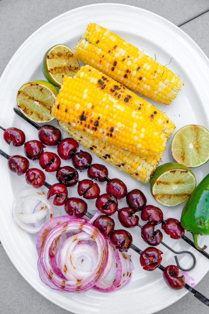 Overhead view of white platter with grilled cherries on skewers, corn on the cob, lime halves, jalapeño, and red onion