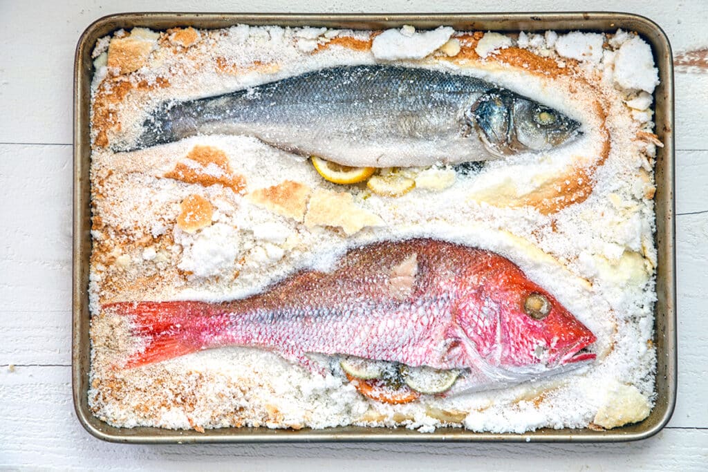 Landscape view of a whole branzino and whole red snapper stuffed with herbs and citrus on a baking sheet with salt chipped away.