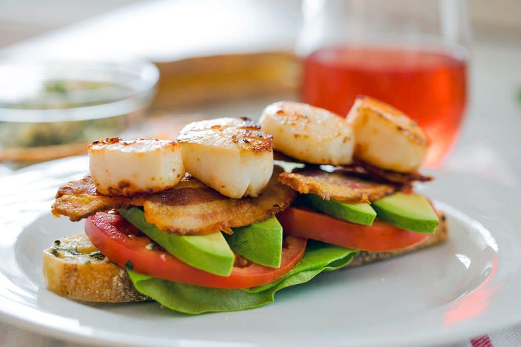Close-up of a seared scallop BLT- a slice of bread with herbed butter, topped with lettuce, tomato slices, avocado slices, crispy bacon, and seared scallops on a white plate