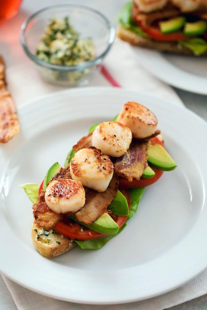 Bird's eye view of seared scallop BLT, crispy bread topped with herbed butter, lettuce, tomatoes, avocado, bacon, and seared scallops on a white plate, with herbed butter in the background