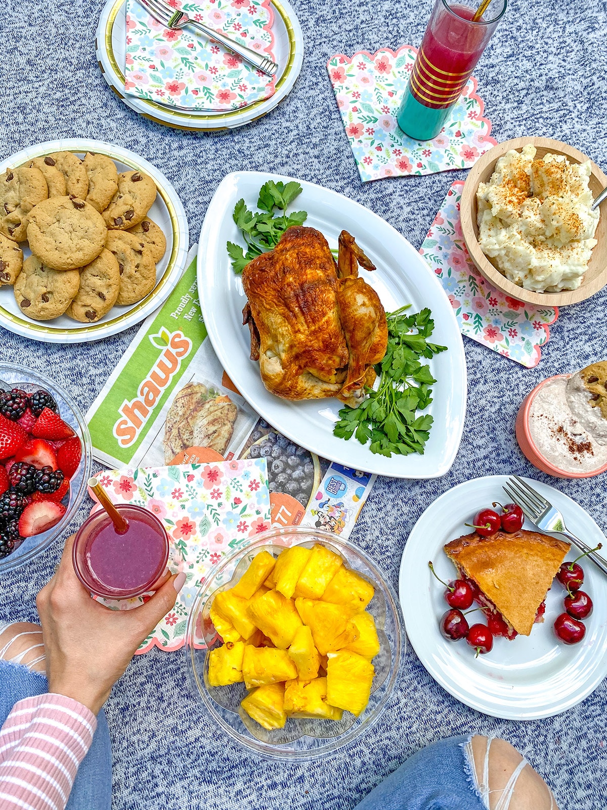 Picnic spread featuring rotisserie chicken, fresh fruit, cookies, cherry pie, potato salad, and juice.