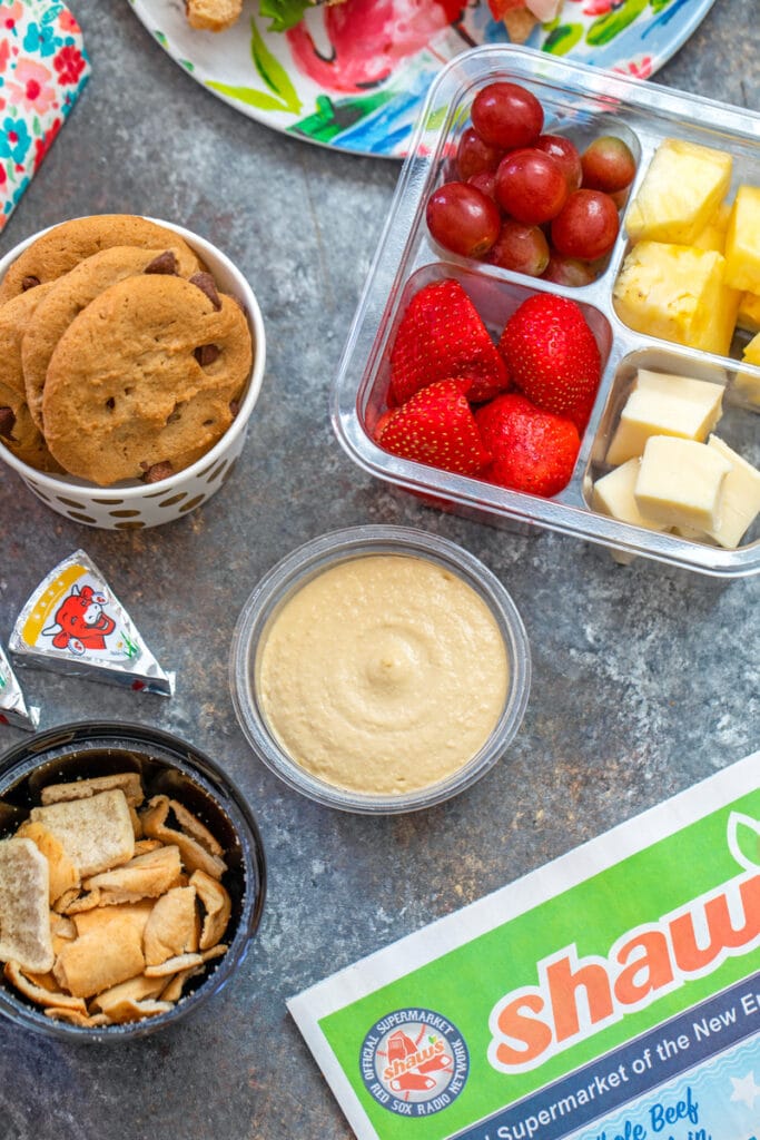 Overhead view of easy back-to-school snacks with hummus, pita chips, fruit, cheese, cookies, etc.