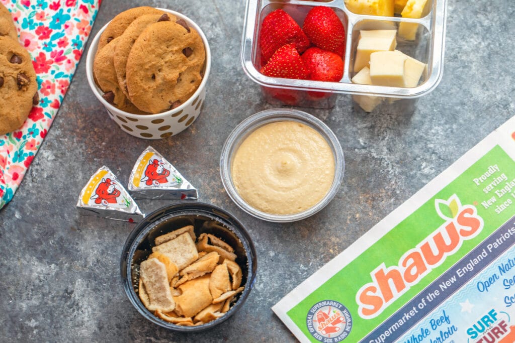 Landscape view of school lunch snacks like hummus, pita chips, cheese, fruit, and cookies