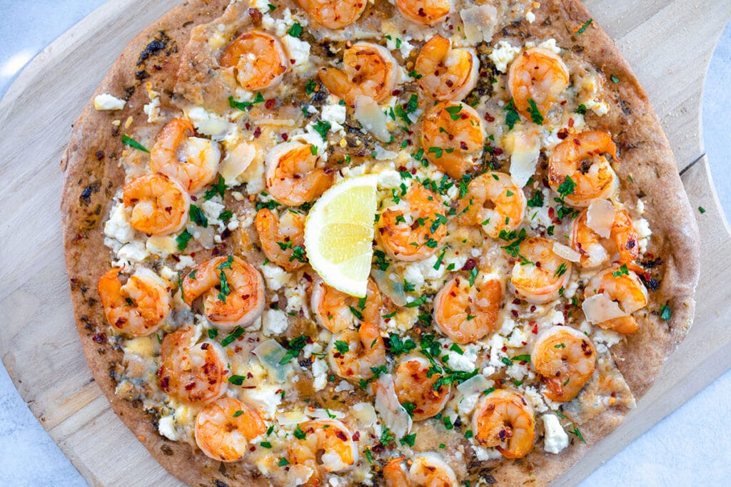 Landscape view of whole shrimp scampi pizza on a wooden board with a lemon in the middle.