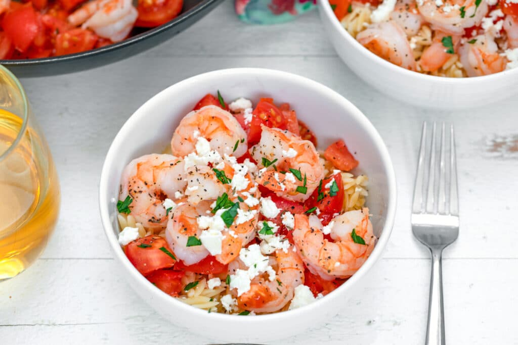 Landscape photo featuring two bowls of orzo with shrimp, feta, tomatoes, and parsley with pan in background and fork and glass of white wine 