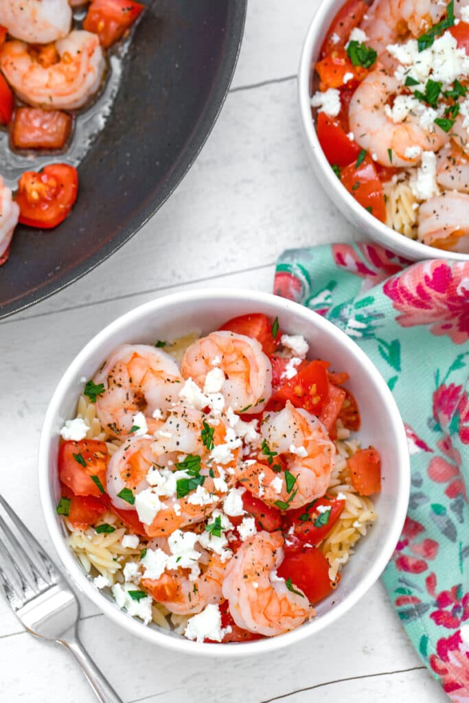 Overhead view of a bowl of orzo topped with shrimp, chopped tomatoes, feta cheese, and parsley with spoon and pretty towel, second bowl of salad and pan with shrimp and tomatoes