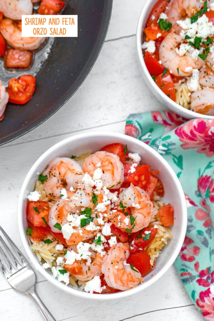 Overhead view of a bowl of orzo topped with shrimp, chopped tomatoes, feta cheese, and parsley with spoon and pretty towel, second bowl of salad and pan with shrimp and tomatoes with recipe title on top