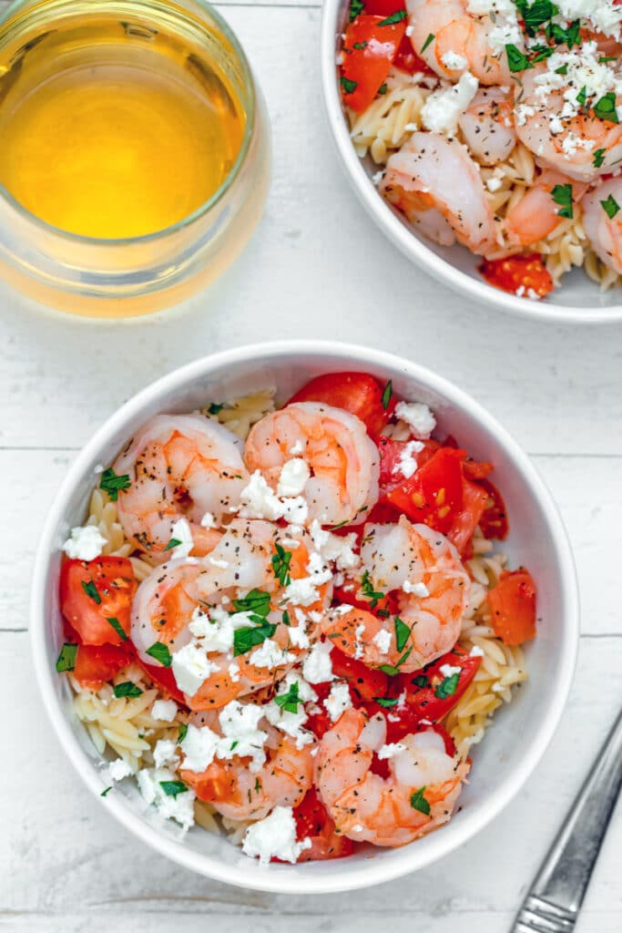 Bird's eye view of a bowl of shrimp and feta orzo salad with tomatoes and parsley with second bowl and glass of white wine in background
