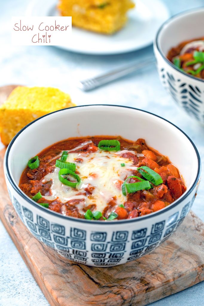 Head-on view of a black and white bowl of slow cooker chili topped with cheese and scallions with second bowl of chili and cornbread in the background with recipe title at top