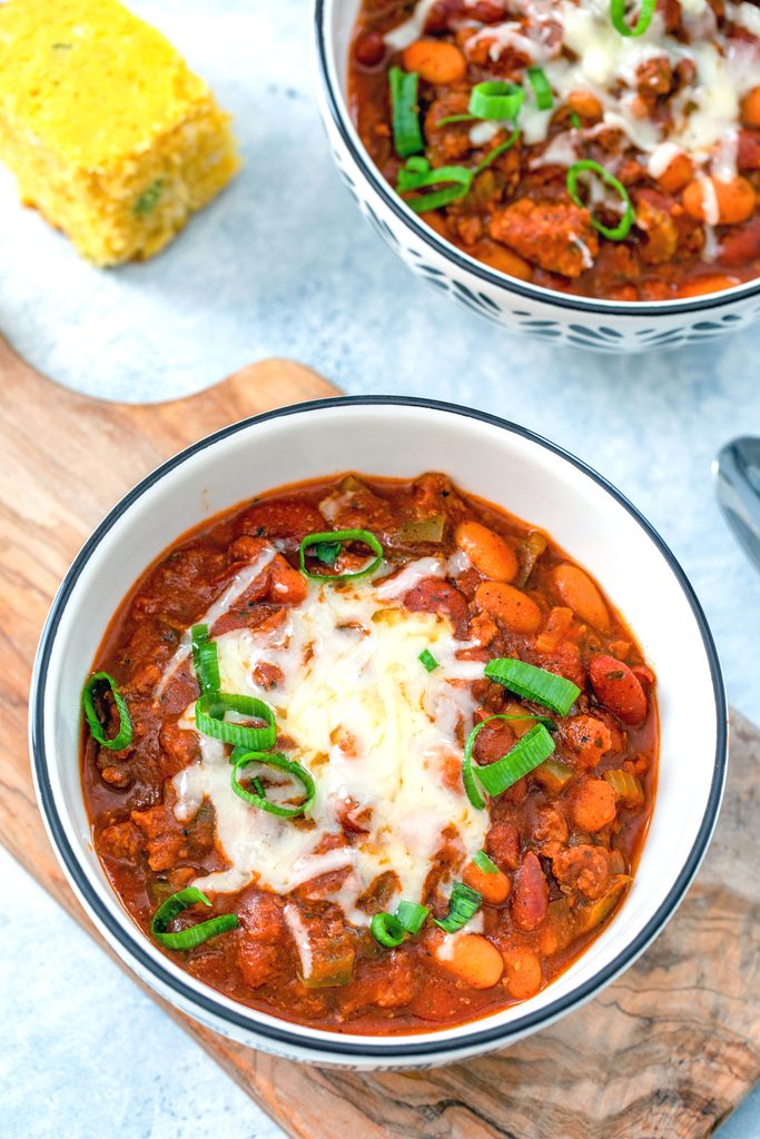 Bird's eye view of slow cooker chili in bowl with cheese and scallions with second bowl of chili and cornbread in background