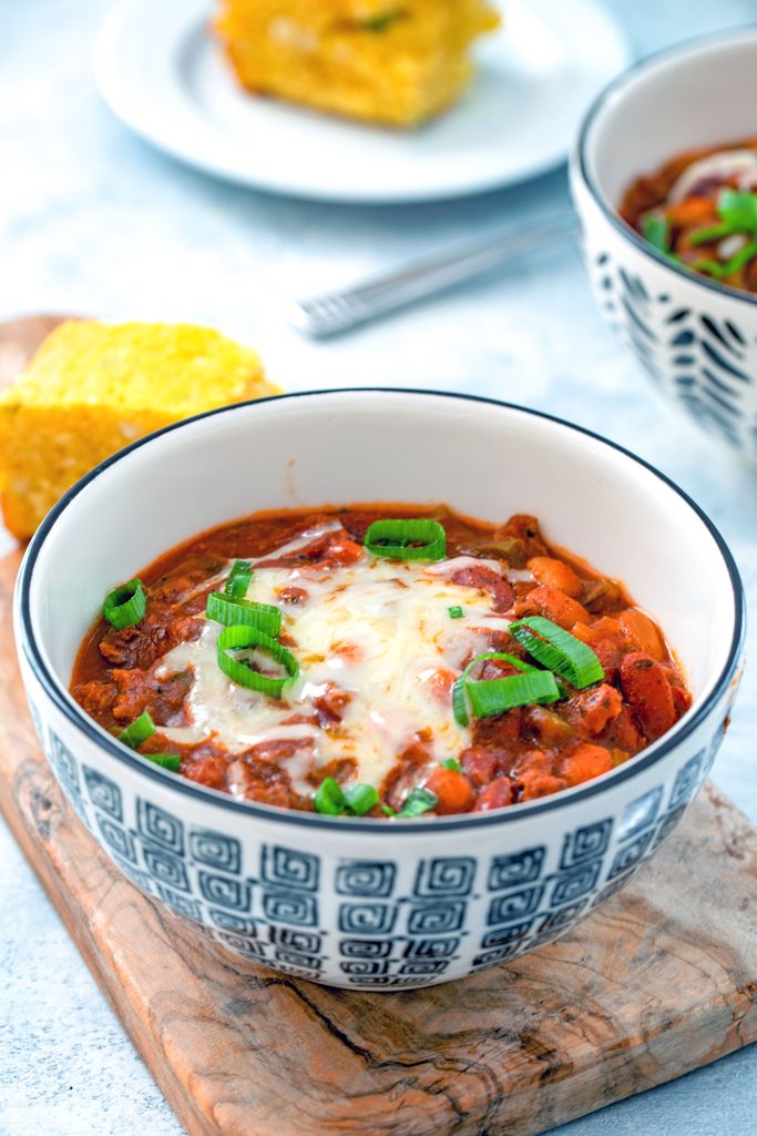 Head-on view of a black and white bowl of slow cooker chili topped with cheese and scallions with second bowl of chili and cornbread in the background