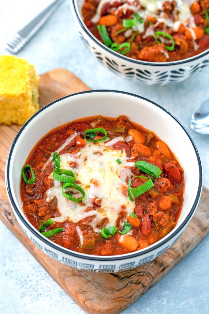 Overhead view of slow cooker chili in a bowl on a wooden board with second bowl of chili and cornbread in the background