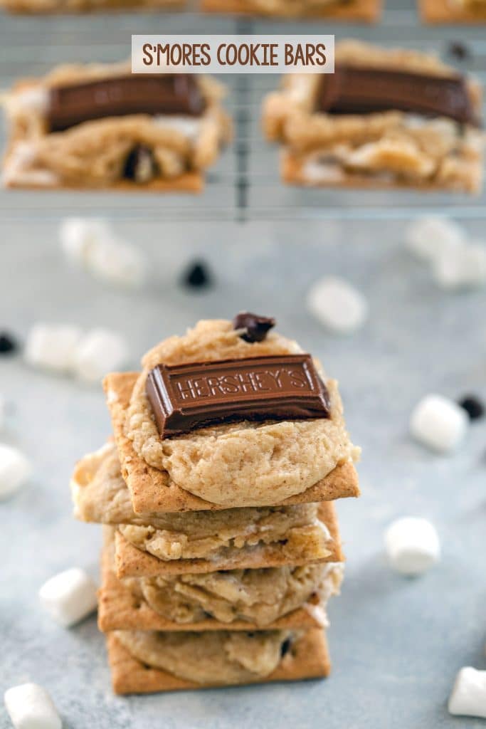 Stack of four s'mores cookie bars surrounded by marshmallows and mini chocolate chips, with a baking rack with more cookies in the background and "S'mores Cookie Bars" text at top