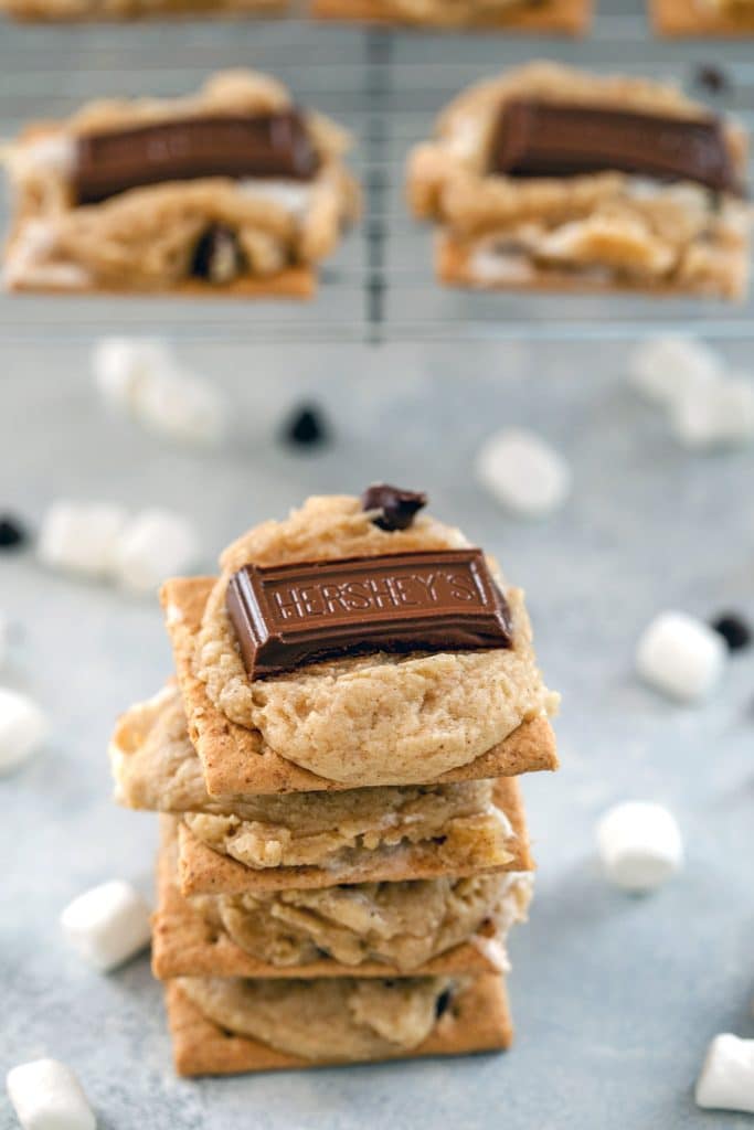 Stack of four s'mores cookie bars surrounded by marshmallows and mini chocolate chips, with a baking rack with more cookies in the background and "S'mores Cookie Bars" text at top