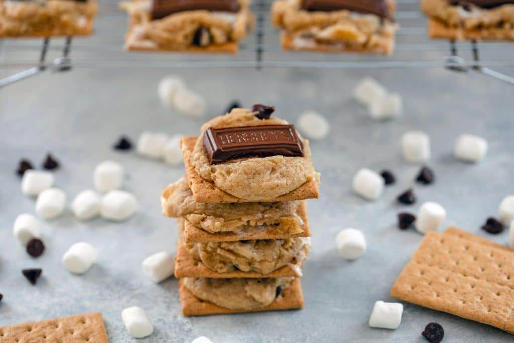 Landscape photo of stack of 4 s'mores cookie bars surrounded by mini marshmallows, chocolate chips, and graham crackers, with baking rack with more cookies in background