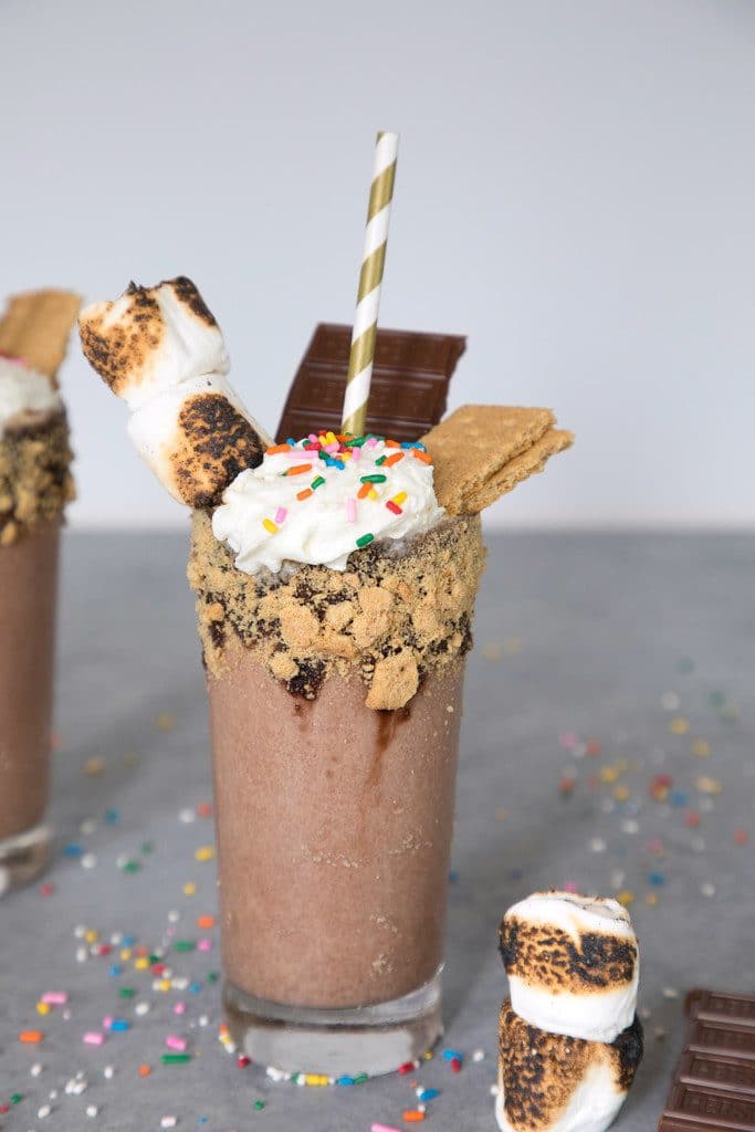 Head-on view of fully loaded bourbon s'mores milkshake with graham rim and toasted marshmallow, chocolate, and graham garnish and sprinkles scattered around