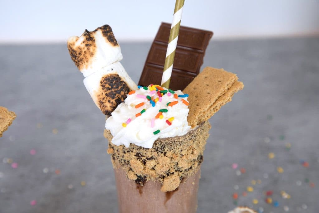 Landscape view of bourbon s'mores milkshake topped with whipped cream, sprinkles, toasted marshmallows, chocolate bars and with a gold striped straw and graham cracker rim