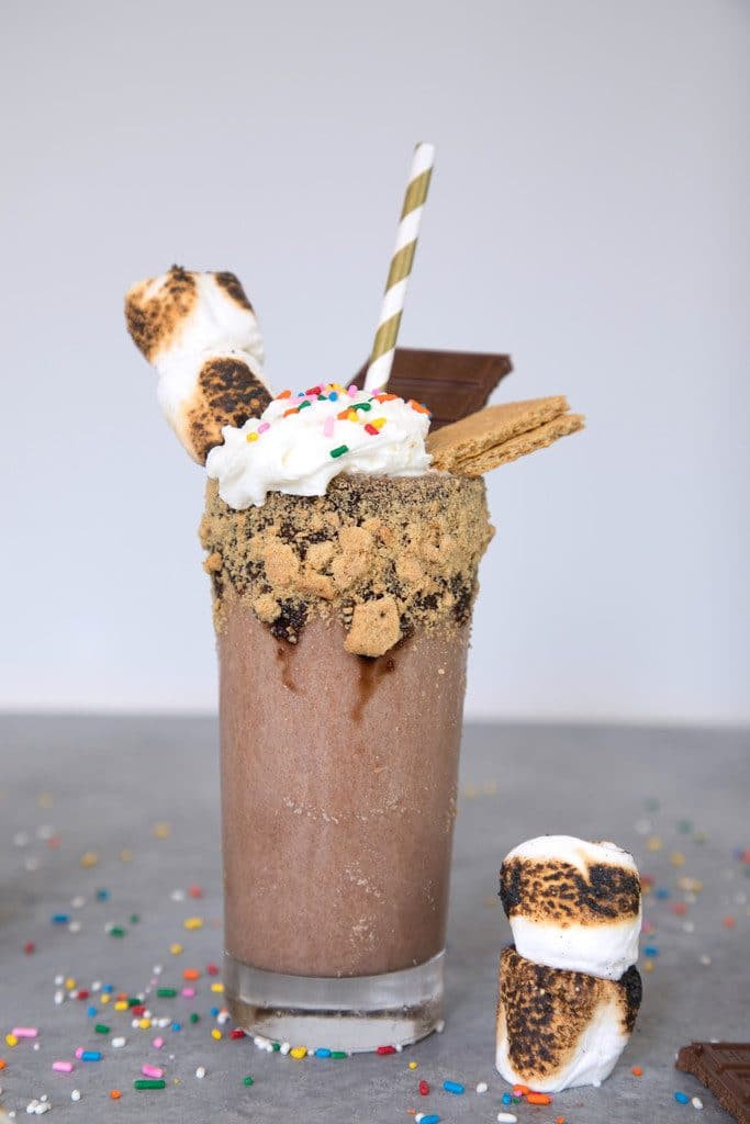 Head on view of bourbon s'mores milkshake on grey background with toasted marshmallows, sprinkles, graham crackers, chocolate bars, and a gold striped straw