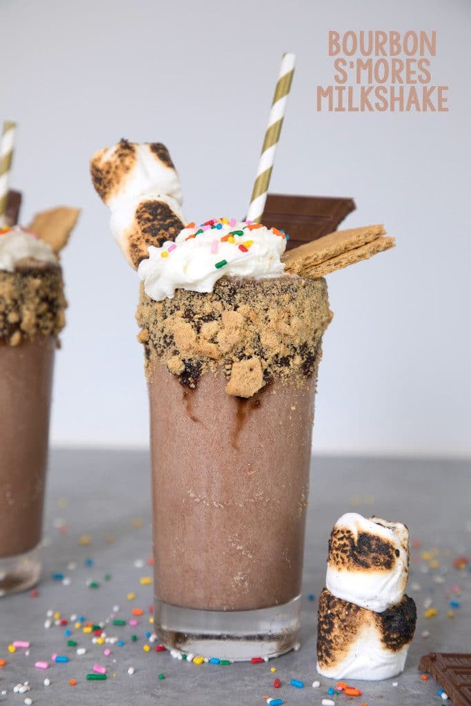 Head-on view of fully loaded bourbon s'mores milkshake with graham rim and toasted marshmallow, chocolate, and graham garnish, sprinkles scattered around, and "Bourbon S'mores Milkshake" text at top
