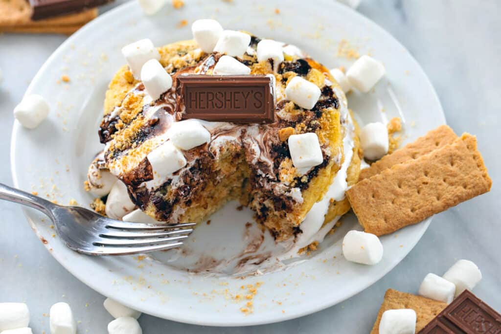 Landscape photo of stack of s'mores pancakes on a white plate with a bite taken out and topped with marshmallow fluff, chocolate sauce, mini marshmallows, chocolate pieces, and graham crackers.