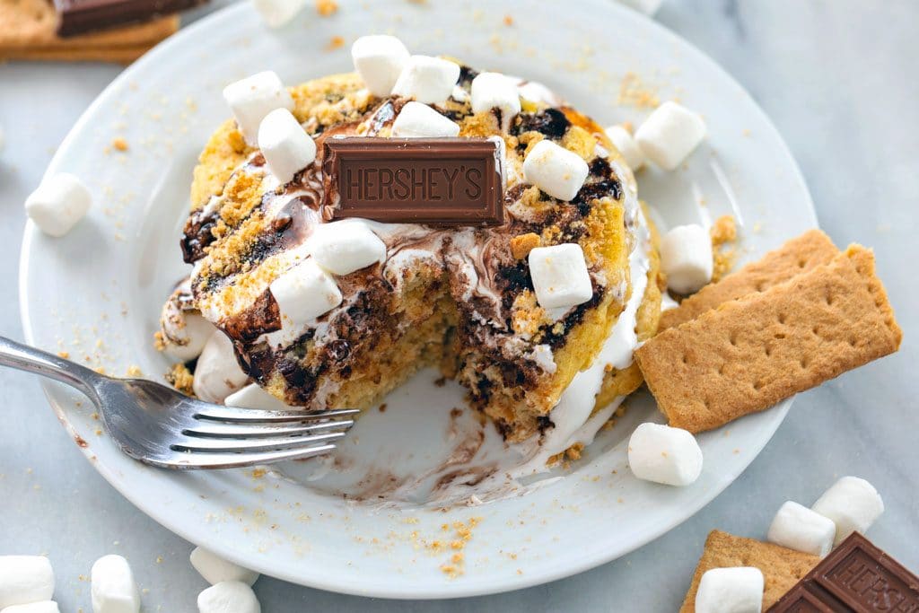 Landscape photo of stack of s'mores pancakes on a white plate with a bite taken out and topped with marshmallow fluff, chocolate sauce, mini marshmallows, chocolate pieces, and graham crackers