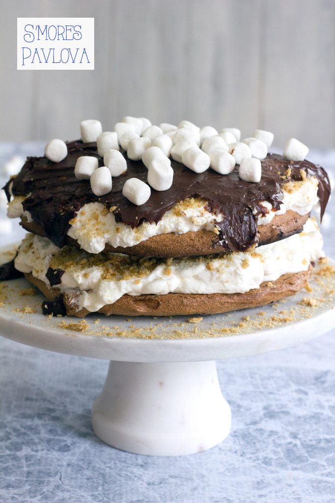 Head-on view of s'mores pavlova cake with whipped cream on a pedestal topped with chocolate ganache, mini marshmallows, and crushed graham crackers with recipe title at top