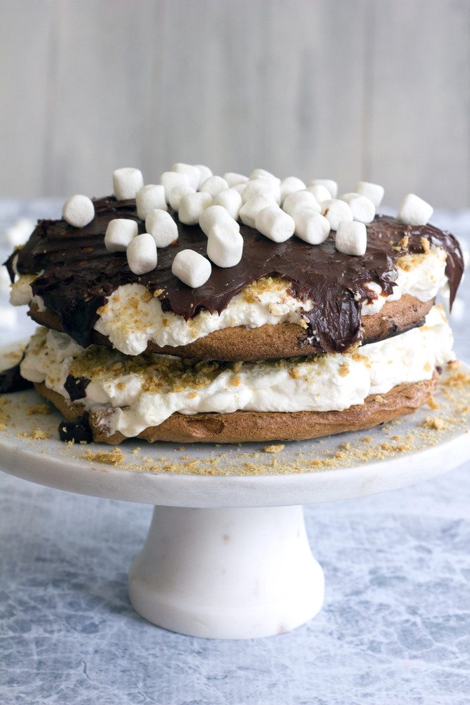 Head-on view of s'mores pavlova cake on a pedestal with whipped cream and topped with chocolate ganache, mini marshmallows, and crushed graham crackers