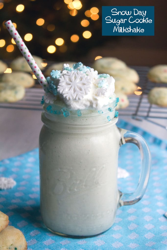 Head-on view of a snow day sugar cookie milkshake in a mason jar with whipped cream and snowflake sprinkles with sugar cookies and twinkling tree lights in the background with recipe title at top