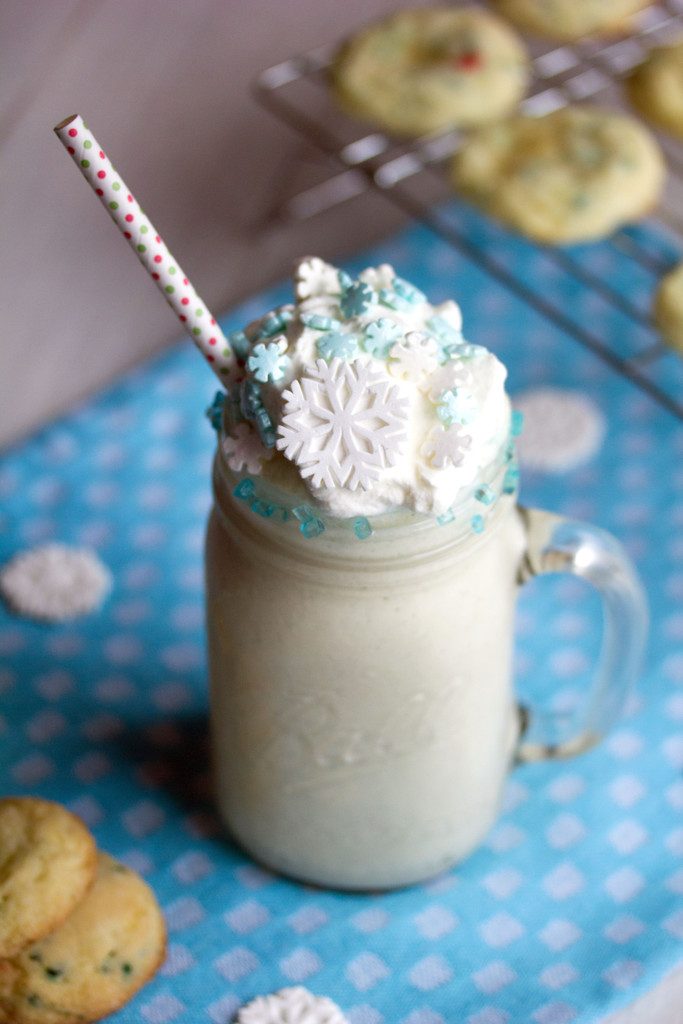 Overhead view of a sugar cookie milkshake in a mason jar with straw, whipped cream, and snowflake sprinkles with sugar cookies in the background