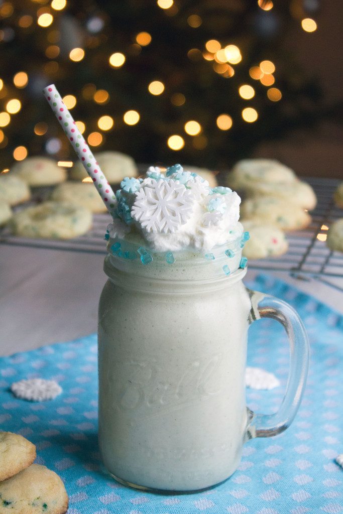 View of a snow day sugar cookie milkshake with whipped cream and snowflake sprinkles with sugar cookies all around and tree with twinkling lights in the background