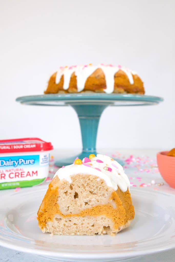 Head-on view of a slice of sour cream pumpkin pie bundt cake with a cake stand with full cake and a sour cream container in the background