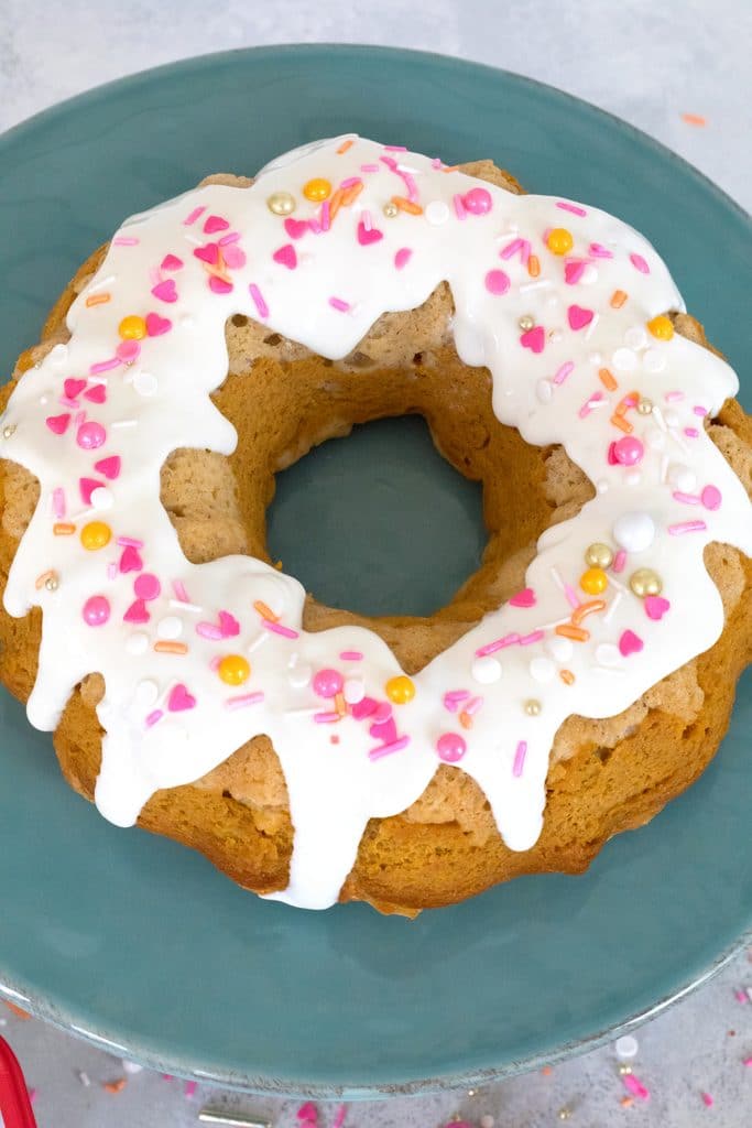 Overhead view of sour cream pumpkin pie bundt cake with white icing and pink and orange sprinkles on a teal cake stand