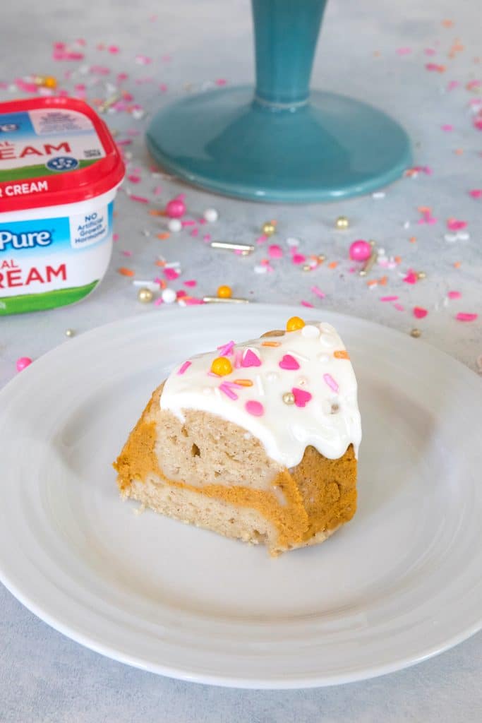 Overhead view of a slice of sour cream pumpkin pie bundt cake with pink and orange sprinkles on a white plate with a sour cream container and cake stand in background