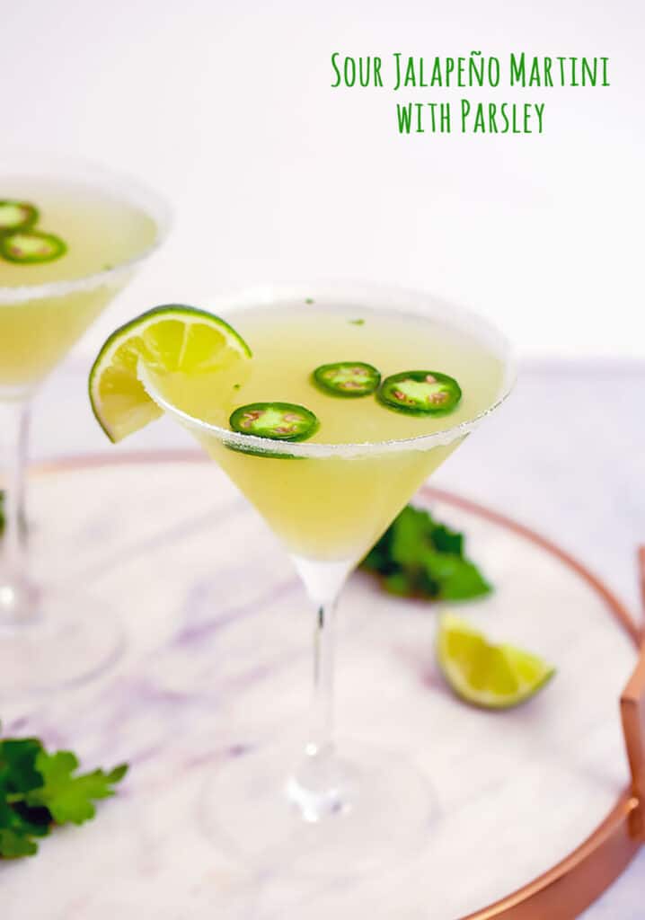 Head-on closeup view of jalapeño martini with vodka, parsley, lemon, and lime juice on a marble tray with lime wedge and parsley in background and recipe title at top