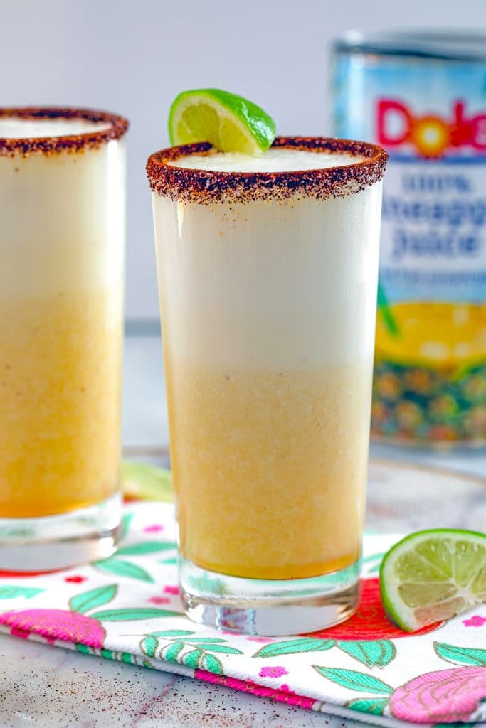 Head-on view of a tall glass of spiced pineapple cocktail with second cocktail, can of pineapple juice, and lime wedge in the background