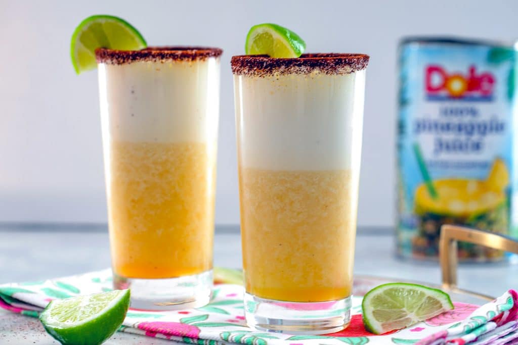 Landscape view of two tall glasses of spiced pineapple cocktail with lime wedges and can of pineapple juice in background