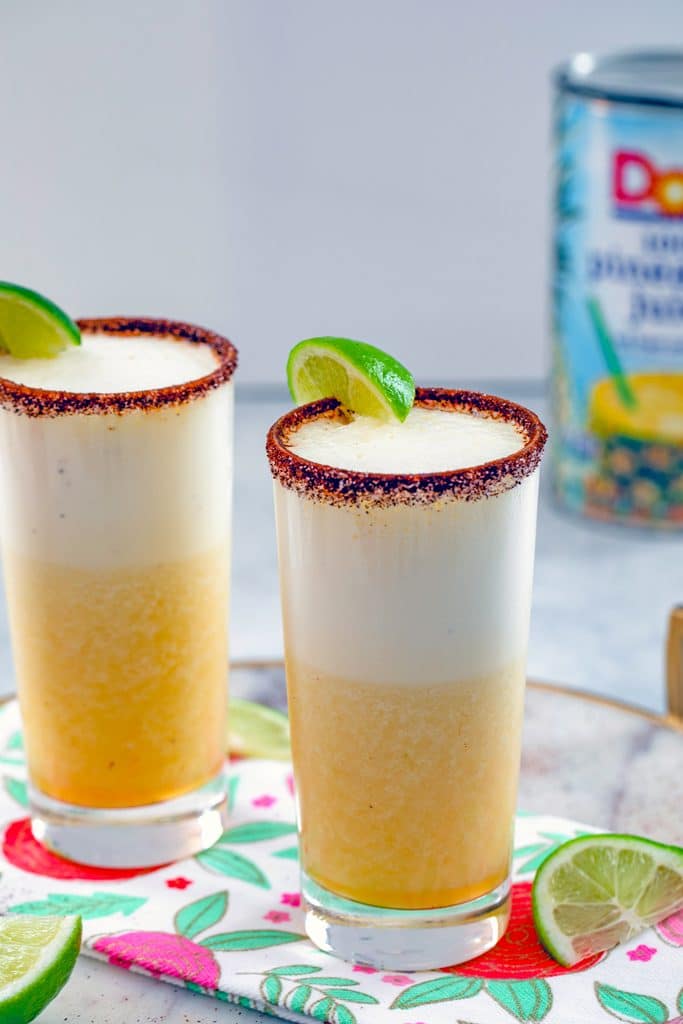 View of two spiced pineapple cocktails with lime wedges and can of pineapple juice in the background