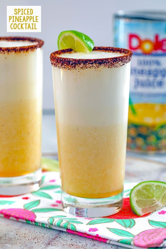 Head-on view of a tall glass of spiced pineapple cocktail with second cocktail, can of pineapple juice, and lime wedge in the background and recipe title at top