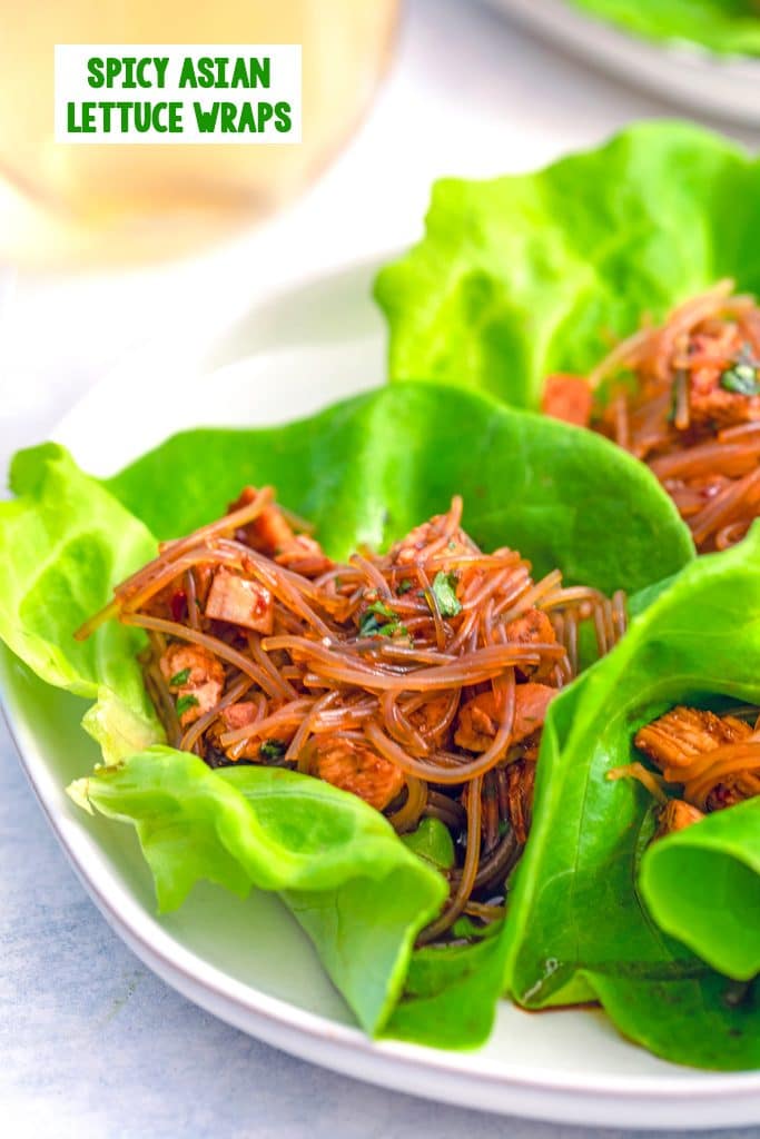 Overhead close-up view of one spicy Asian lettuce wrap on a plate with two other lettuce wraps surrounding it, glass of white wine in the background, and recipe title at top