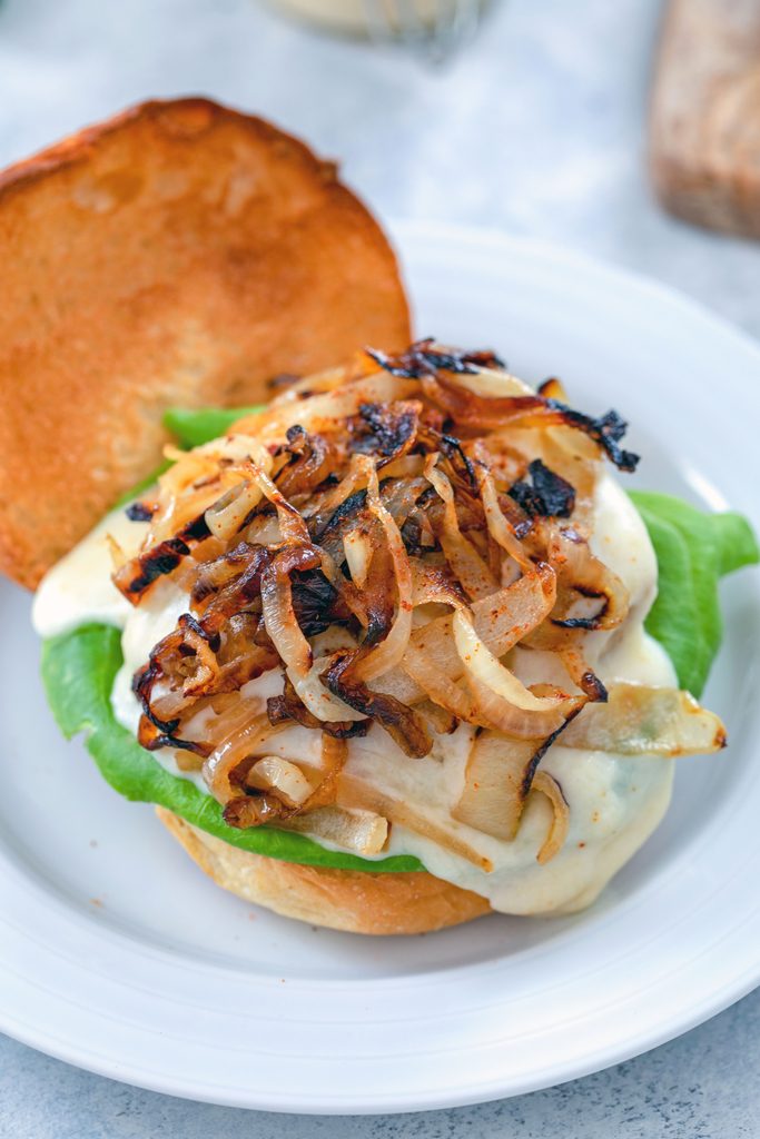 Overhead view of open-faced spicy cheesy chicken burger on brioche roll topped with lettuce, cheese sauce, and caramelized onions