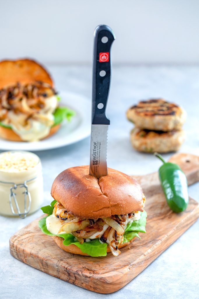 Head-on view of a spicy cheesy chicken burger on a small wood cutting board with a knife stuck in it with jalapeño, jar of cheese sauce, second burger, and more chicken burger patties in background